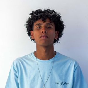 Casual portrait of a young man with curly hair wearing a blue t-shirt and necklace.