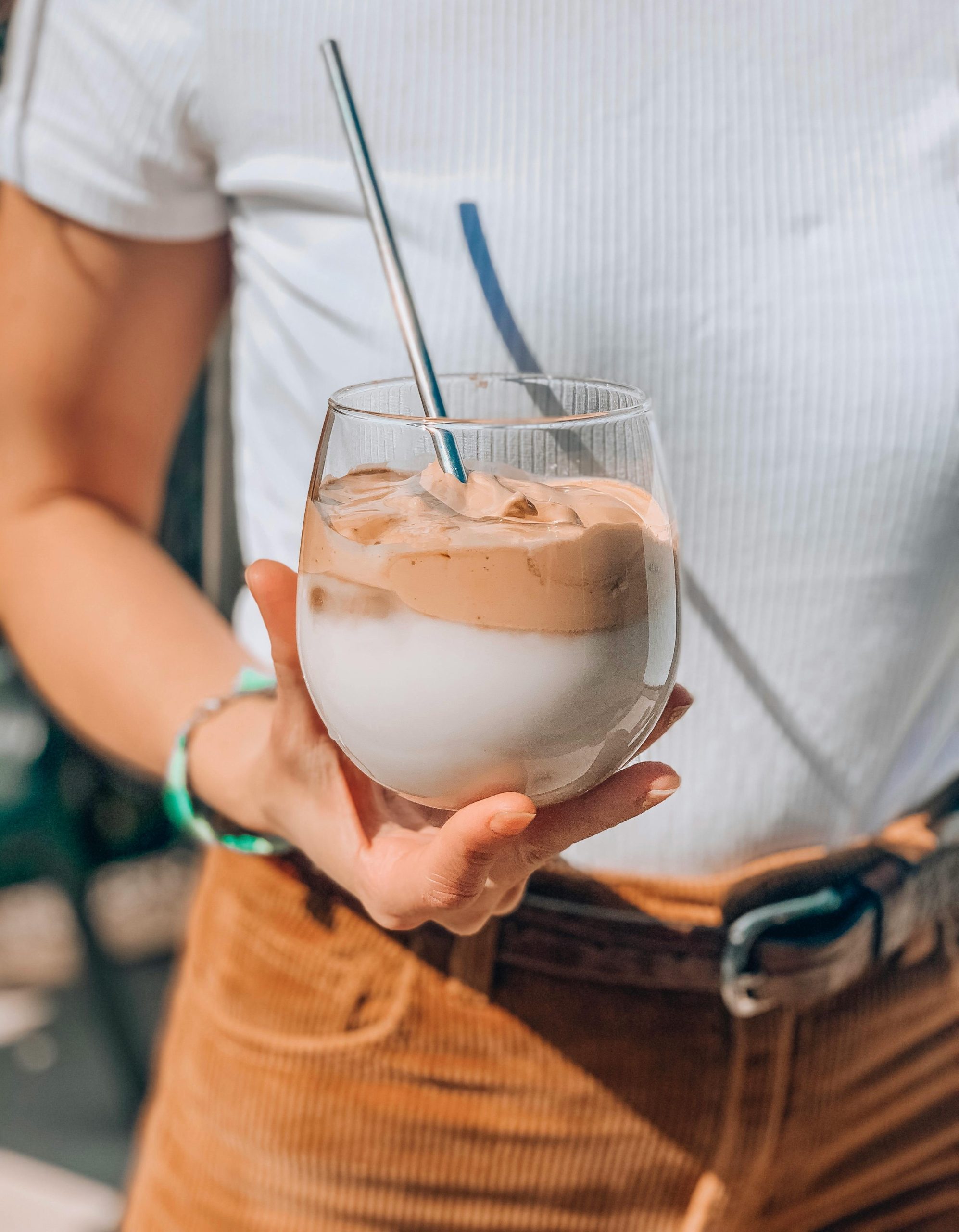 A close-up of a woman holding a glass of creamy Dalgona coffee with a stainless steel straw.