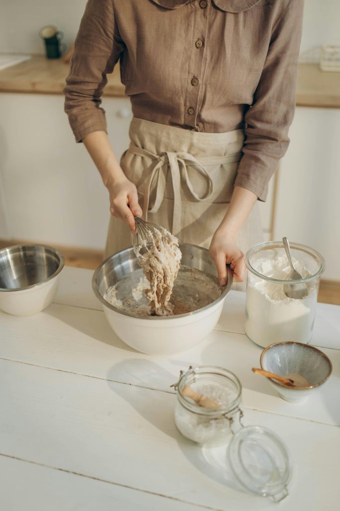 Woman mixing dough with a whisk in the kitchen, surrounded by baking ingredients.
